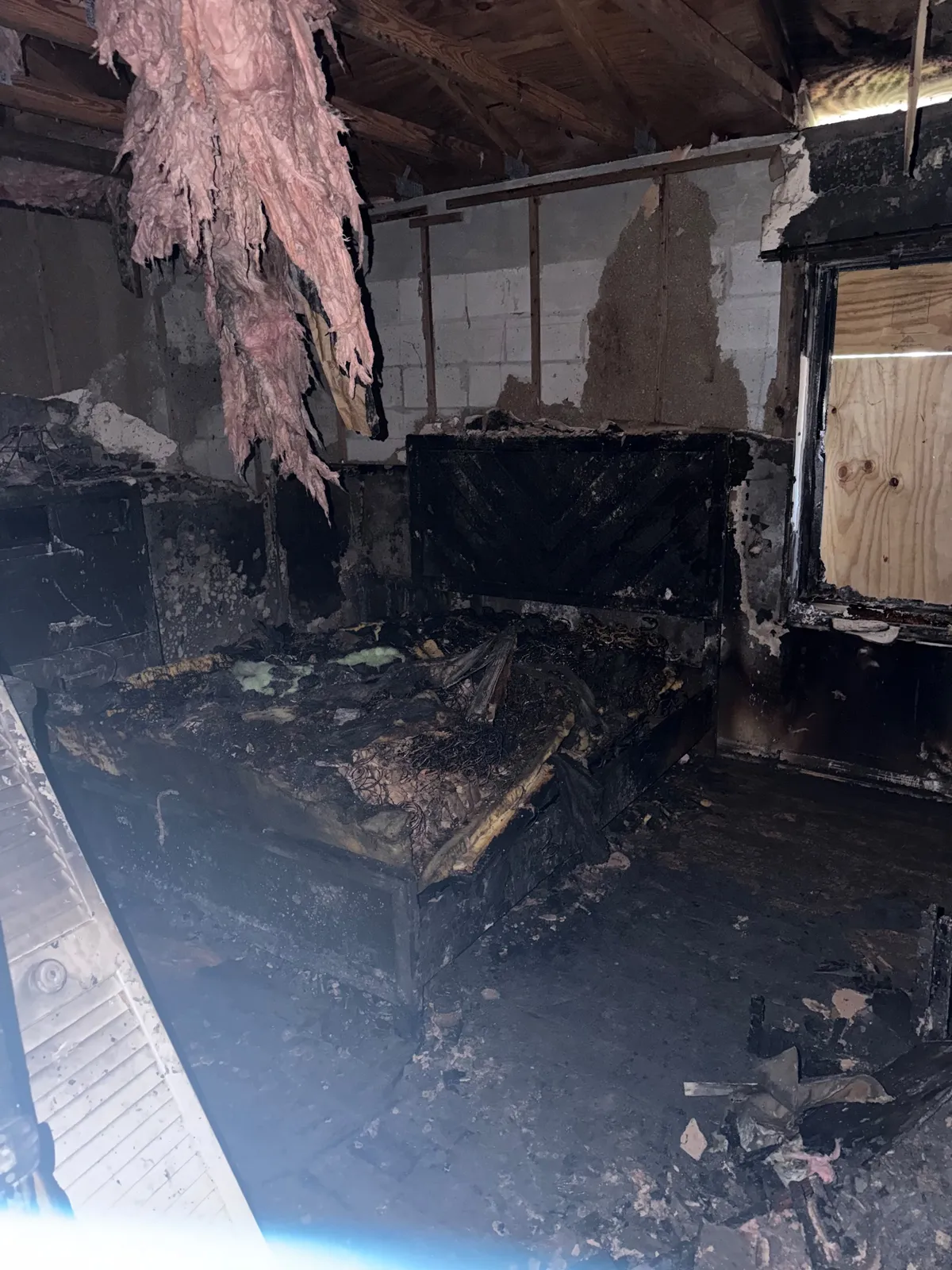Fire-damaged bedroom with a charred bed frame, burned insulation and a boarded window.