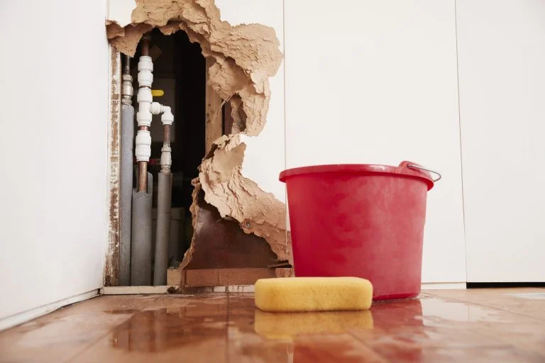 Water-damaged wall opened to a burst pipe with a cleanup bucket and sponge on the floor.
